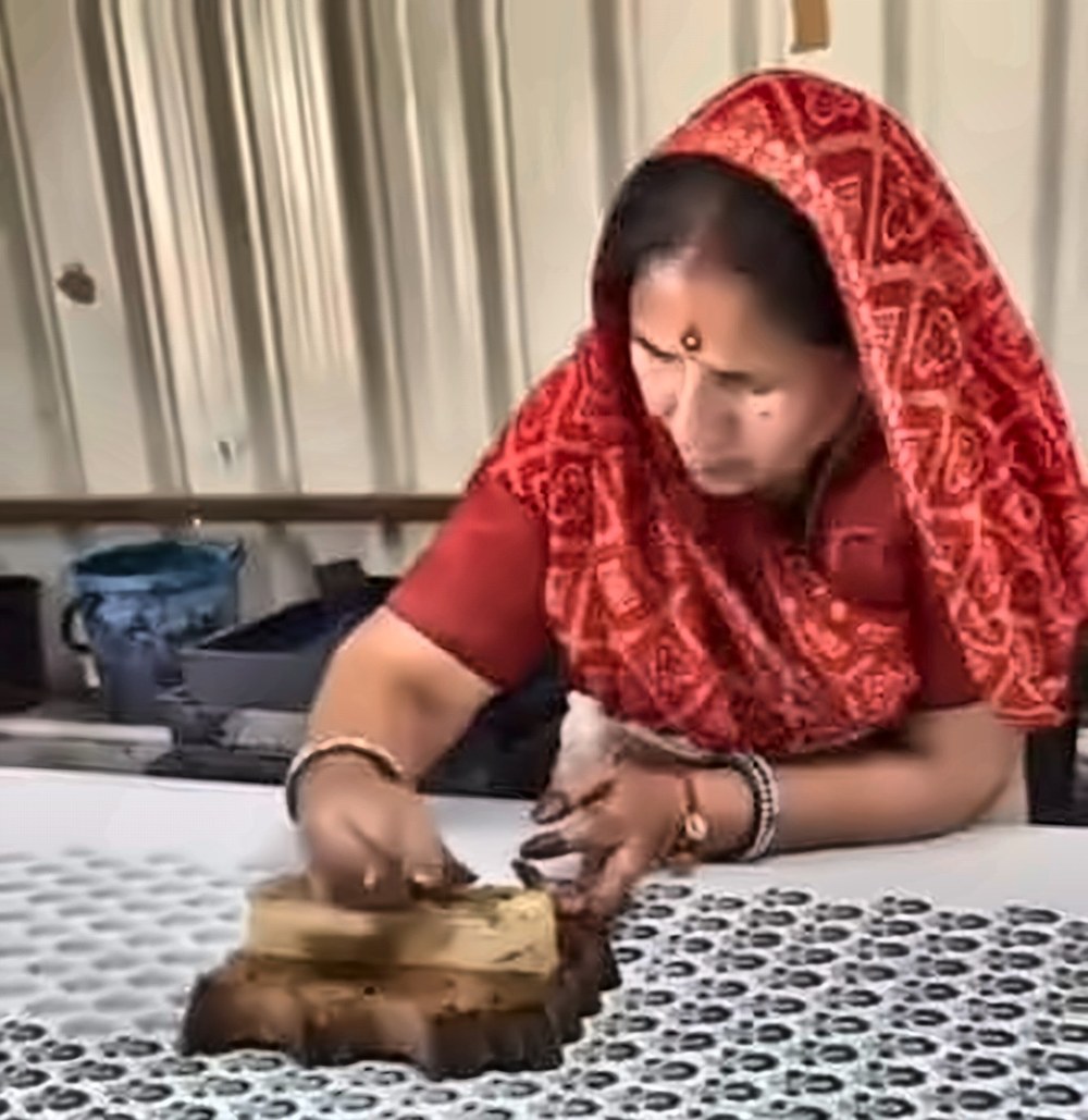 An artisan in a red bandhani sari pressing a carved wooden block onto white cotton